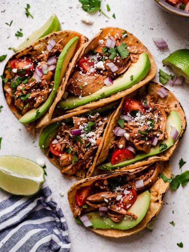 Overhead view of a white countertop with chicken tacos lined up in a row topped with fresh cilantro and red onion. 