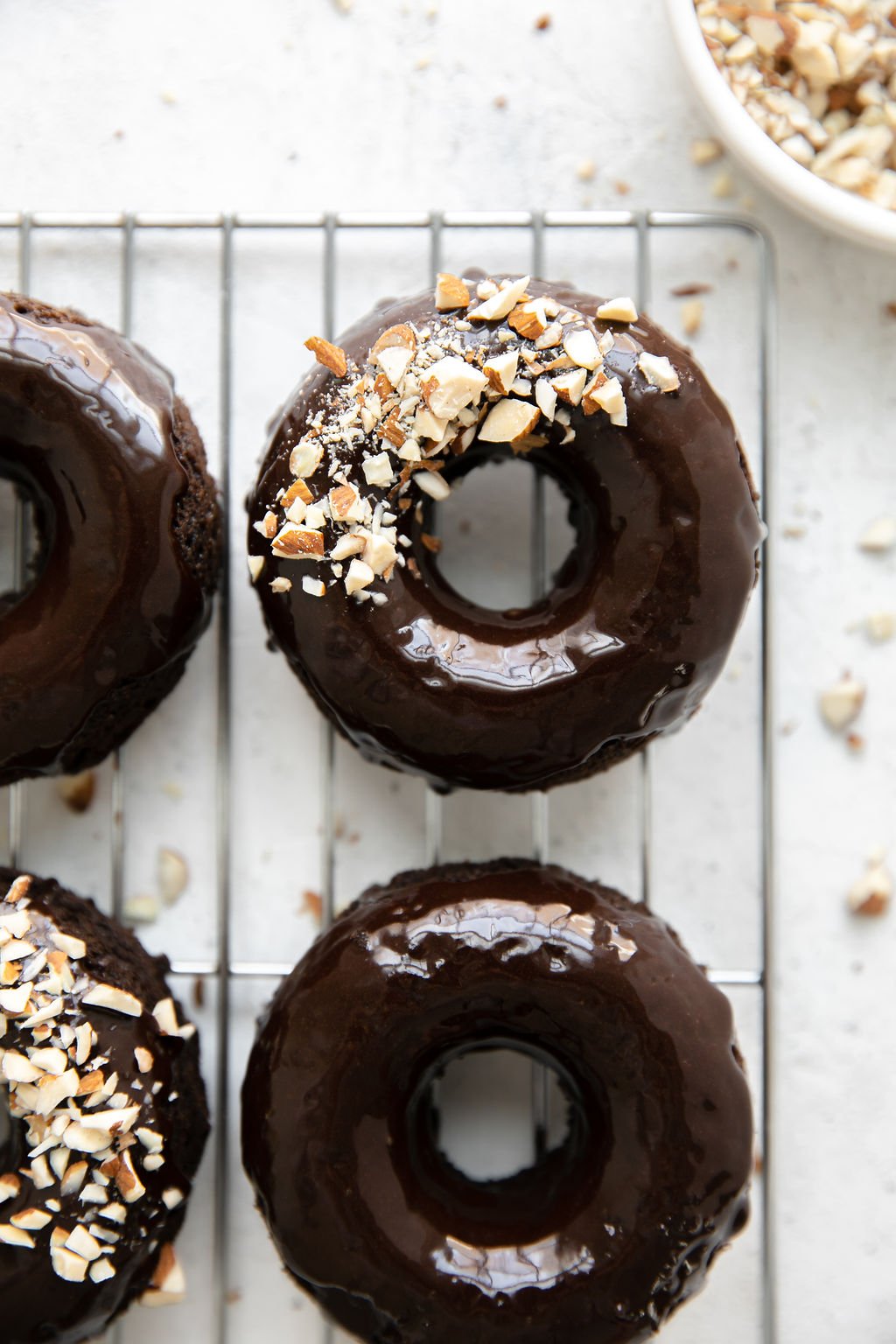 Overhead view of flourless chocolate donuts with shiny chocolate icing and crushed almonds on one side of the donut, lined up on a wiring cooling rack.