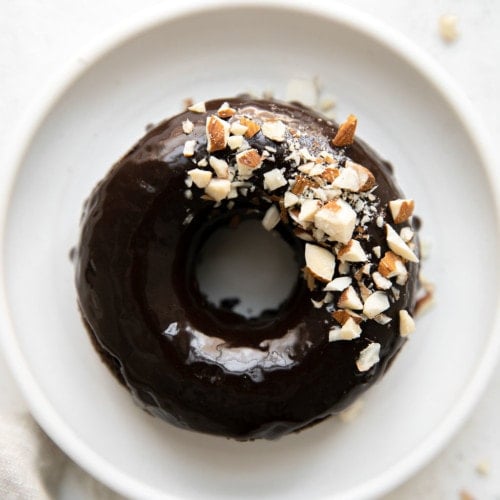 Overhead view of flourless chocolate donut with chocolate icing and crushed almonds on a small white plate.