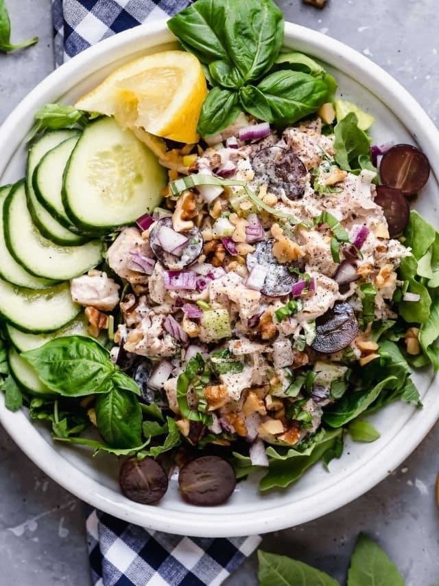 Chicken Waldorf Salad on a white plate over a bed of mixed greens topped with walnuts and fresh herbs.