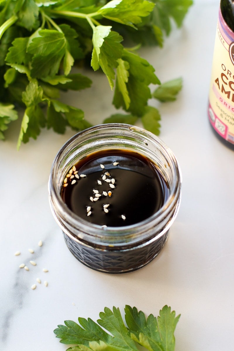 A short glass jar filled with coconut aminos a few sesame seeds sprinkled on top of dark liquid.