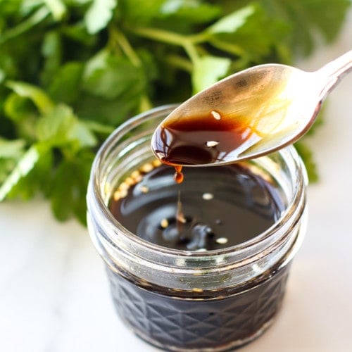 Coconut aminos in a short glass jar showing dark liquid, a spoon taking a small serving out.