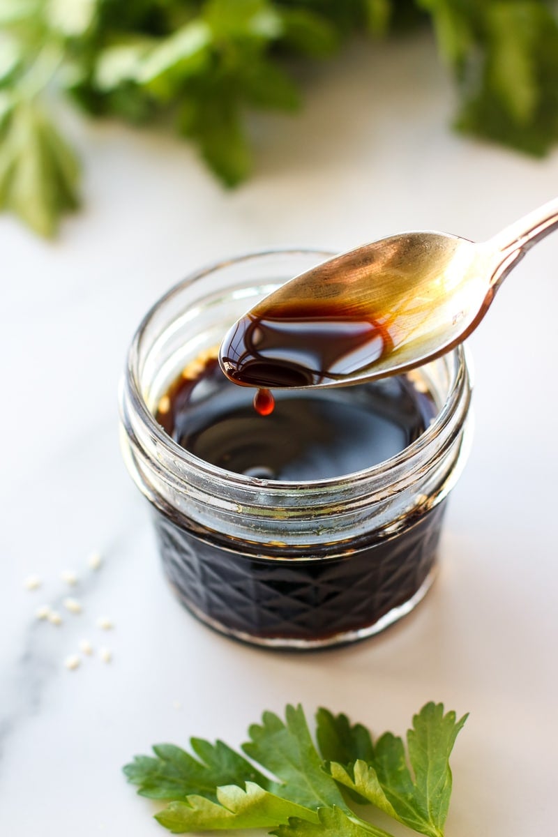 A short glass jar filled with coconut aminos, a spoon taking out a small scoop, a drop of coconut aminos dripping off the bottom of the spoon.
