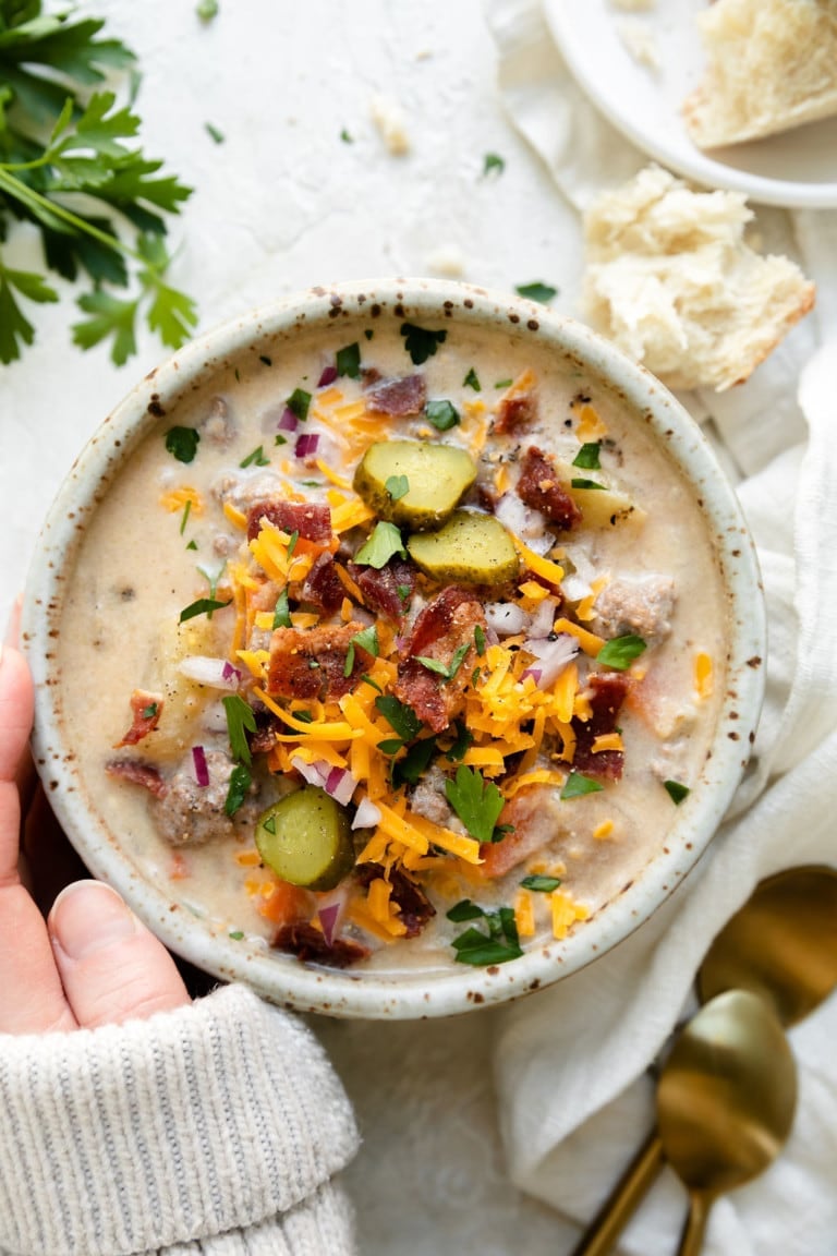 White bowl with cheeseburger soup and topped with bacon, cheese, and pickles. 