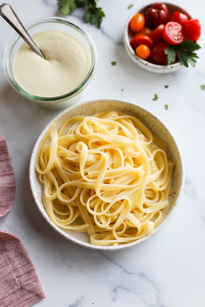 A bowl of pasta next to a bowl of white creamy Vegan Alfredo Sauce