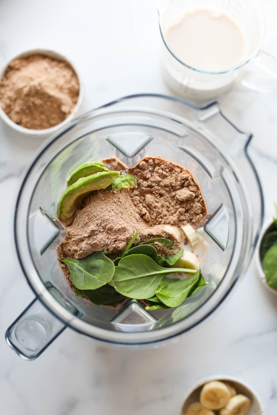 Overhead view looking down into a high speed blender cup filled halfway with fruit, spinach, avocado, and chocolate protein powder. 