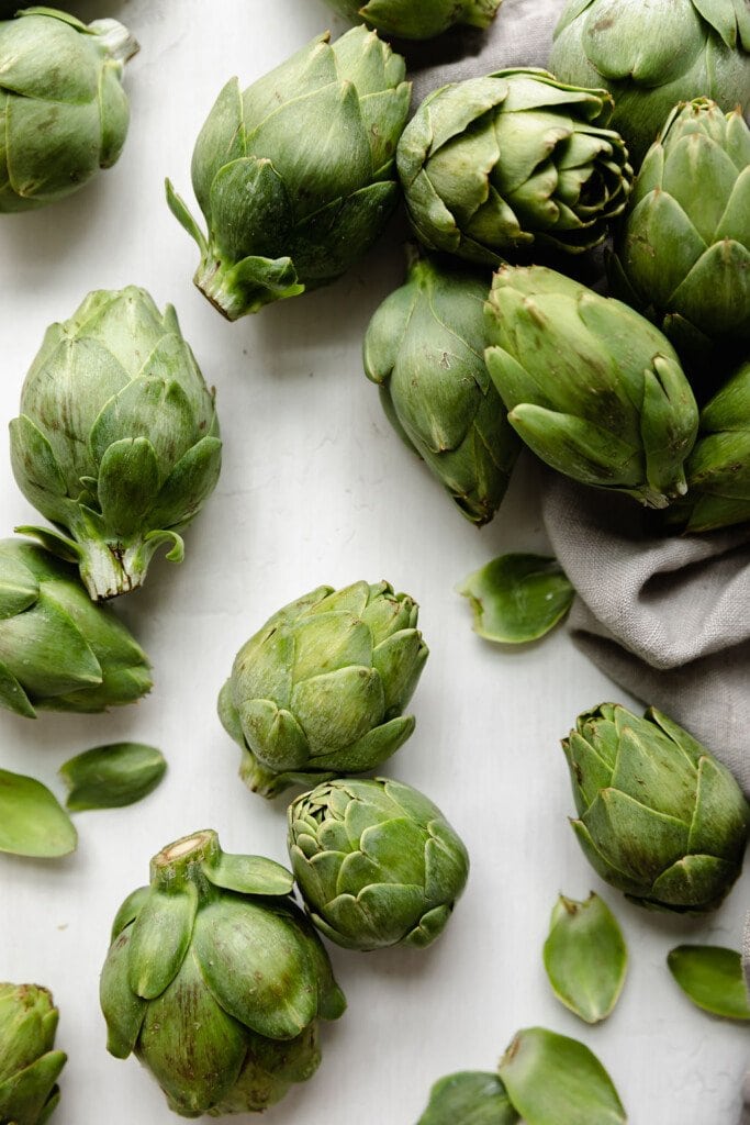 Several baby artichokes arranged together on a white background with a light tan dishcloth to one side.