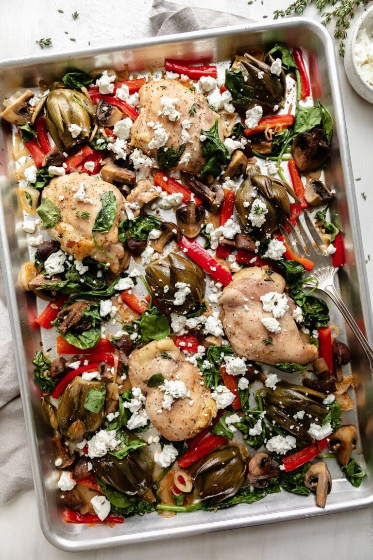 Overhead view of a sheet pan filled with chicken breasts alongside baby artichokes, mushrooms, and bell peppers topped with goat cheese.