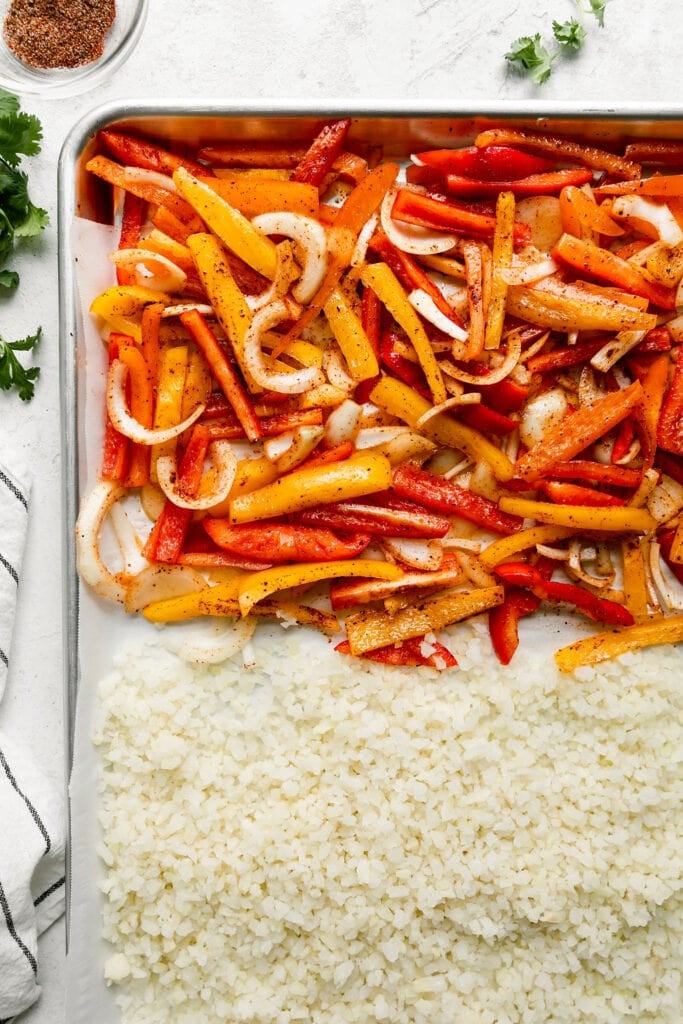 Overhead view of sheet pan half filled with fajita seasoned bell peppers and onions and half filled with cauliflower rice.