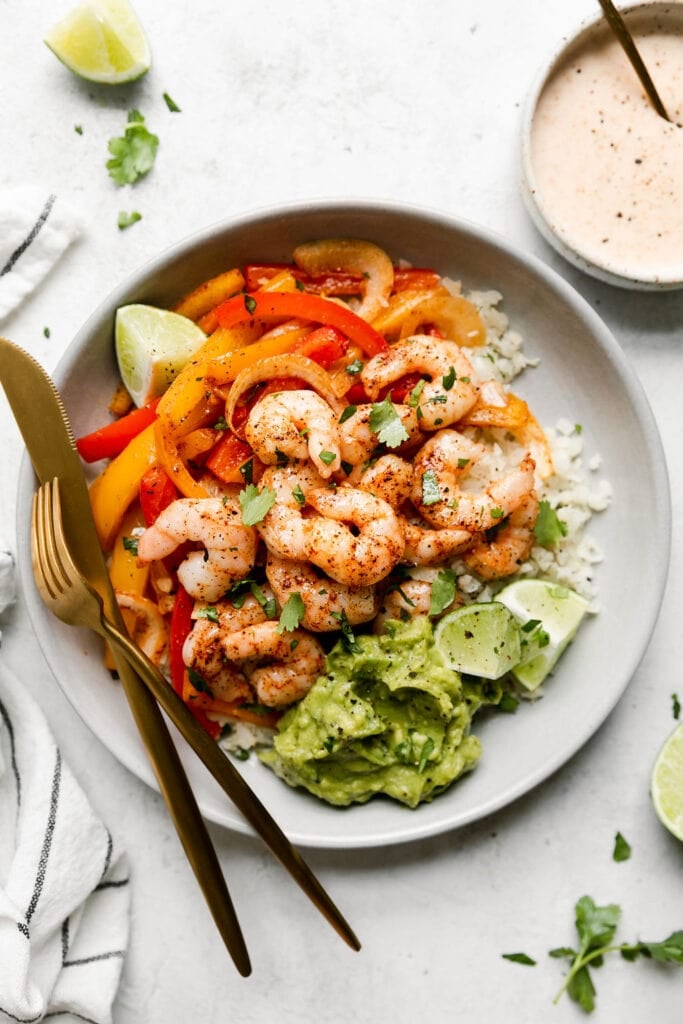 Overhead view of sheet pan shrimp fajita bowl served in a shallow bowl topped with avocado, lime wedges, and fresh cilantro.