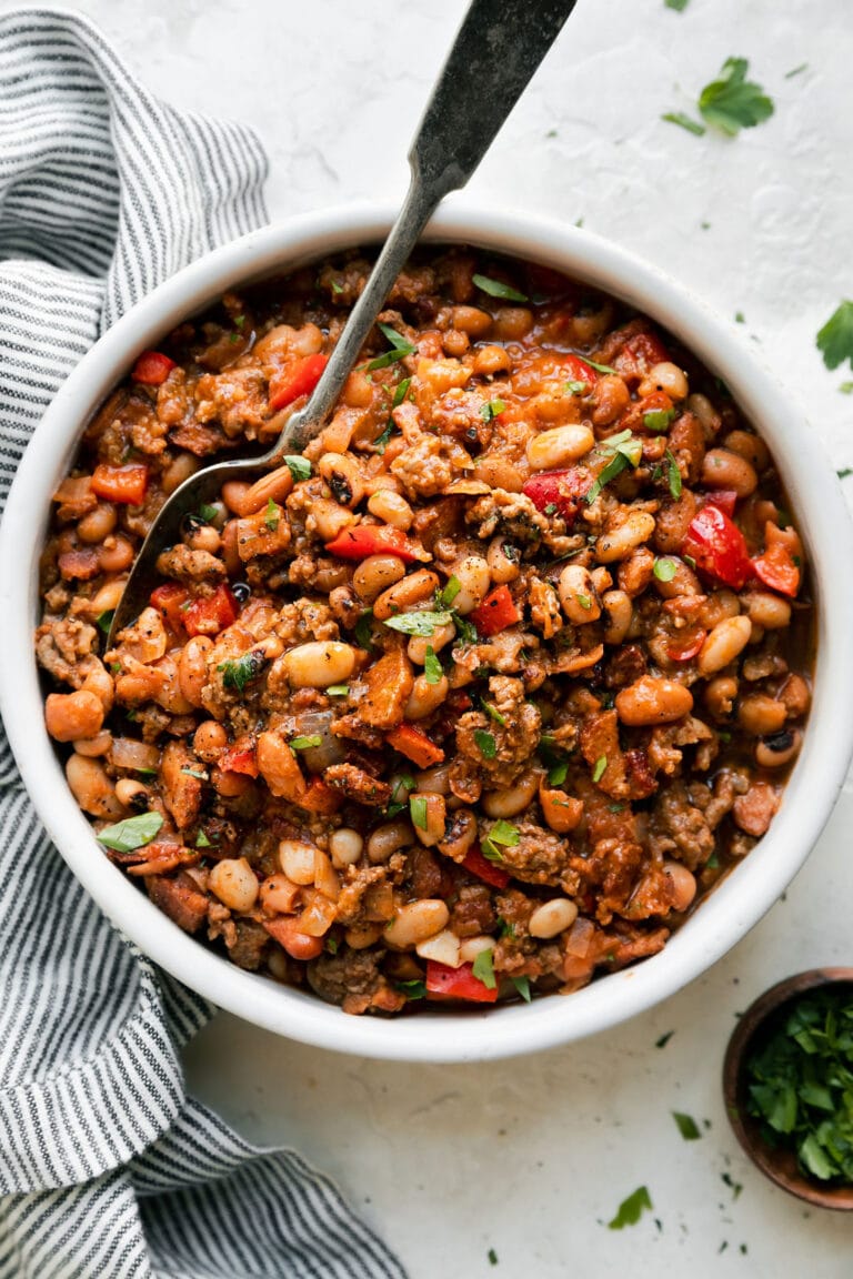 White bowl filled with slow cooker cowboy beans and sauce being scooped with a metal spoon. 