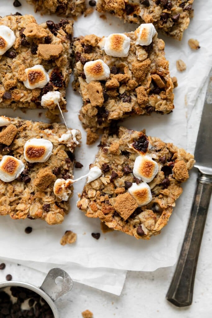 Overhead close up view of chewy s'mores granola bars topped with toasted mini marshmallows, melty mini chocolate chips, and broken up graham crackers, cut into squares on parchment paper.
