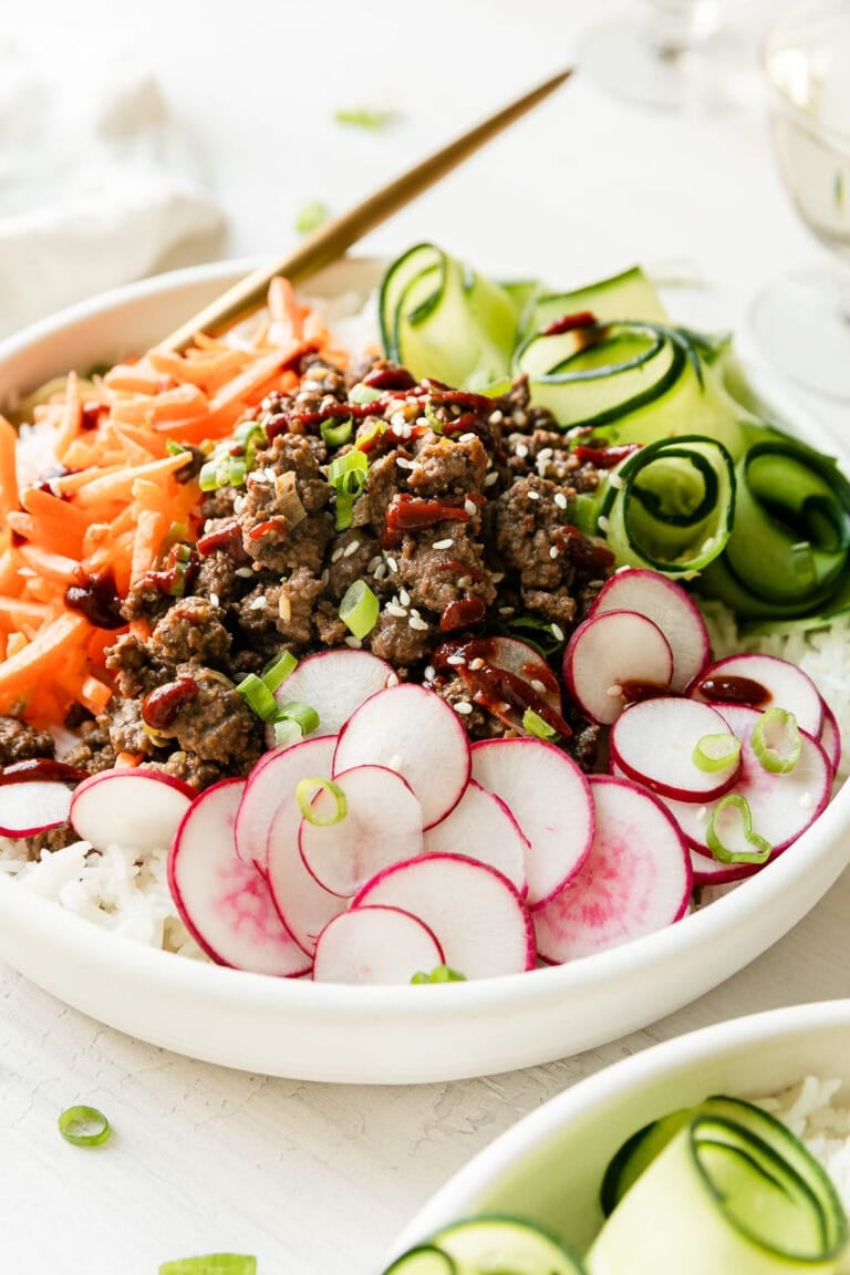 Close up view of a white bowl filled with rice, radish slices, carrot matchsticks, cucumber ribbons, and Korean ground beef topped with green onions and sesame seeds. 