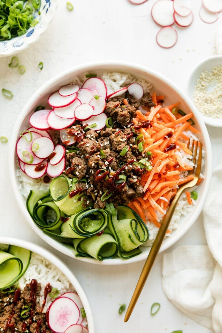 Overhead view of a Korean ground beef bowl with shredded carrots, curled thin slices of cucumber, and sliced radishes served over rice in white bowl.