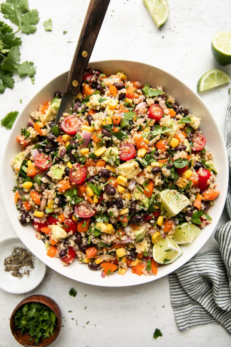 Overhead view Southwest flavored quinoa salad with corn, avocado, tomatoes, and lime slices, in a white serving bowl with wooden serving spoon.