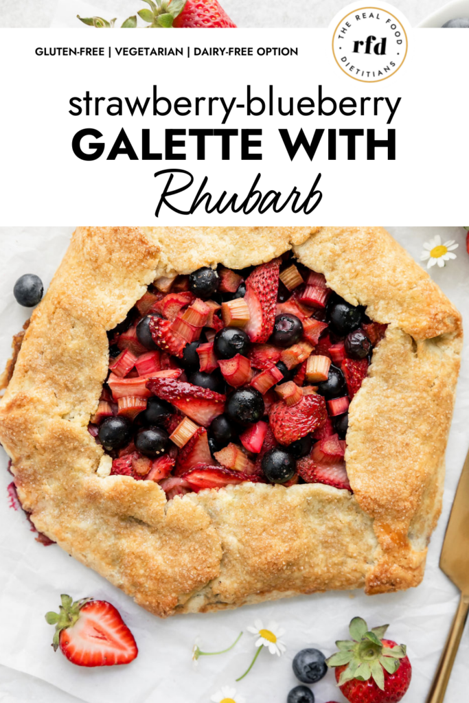 Overhead view strawberry blueberry galette with rhubarb on parchment paper, fresh berries and Shasta daisies scattered around Galette.