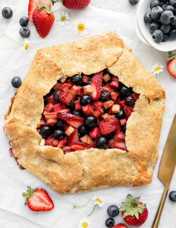 Overhead view strawberry blueberry galette with rhubarb in center of parchment paper, fresh berries and Shasta daisies scattered around.