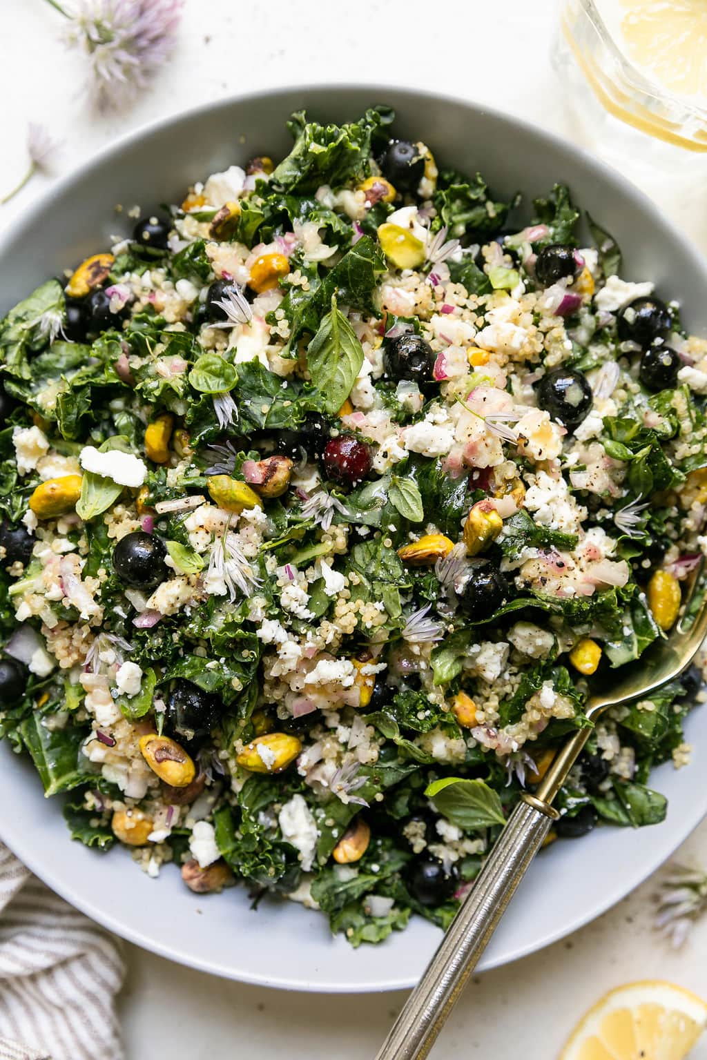 Overhead view quinoa kale salad with blueberries, feta, and pistachios served in white bowl with gold fork.