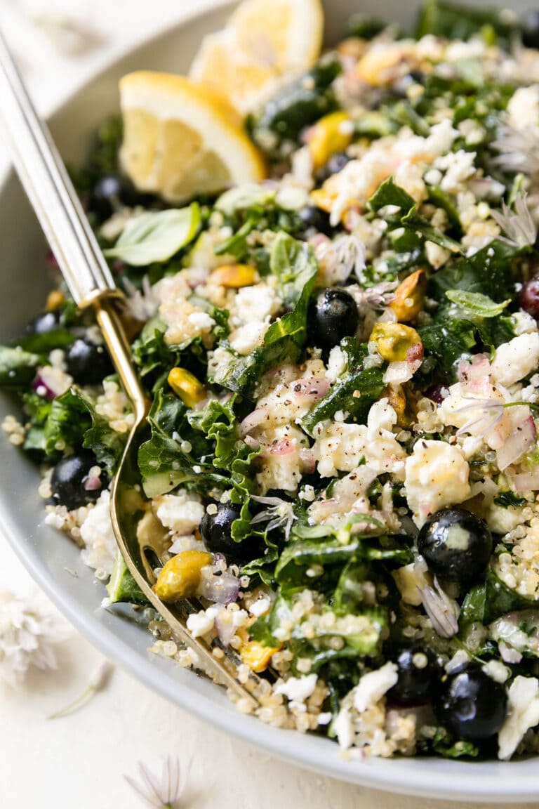 Close up view kale and quinoa salad with blueberries in a serving bowl garnished with lemon wedges. 