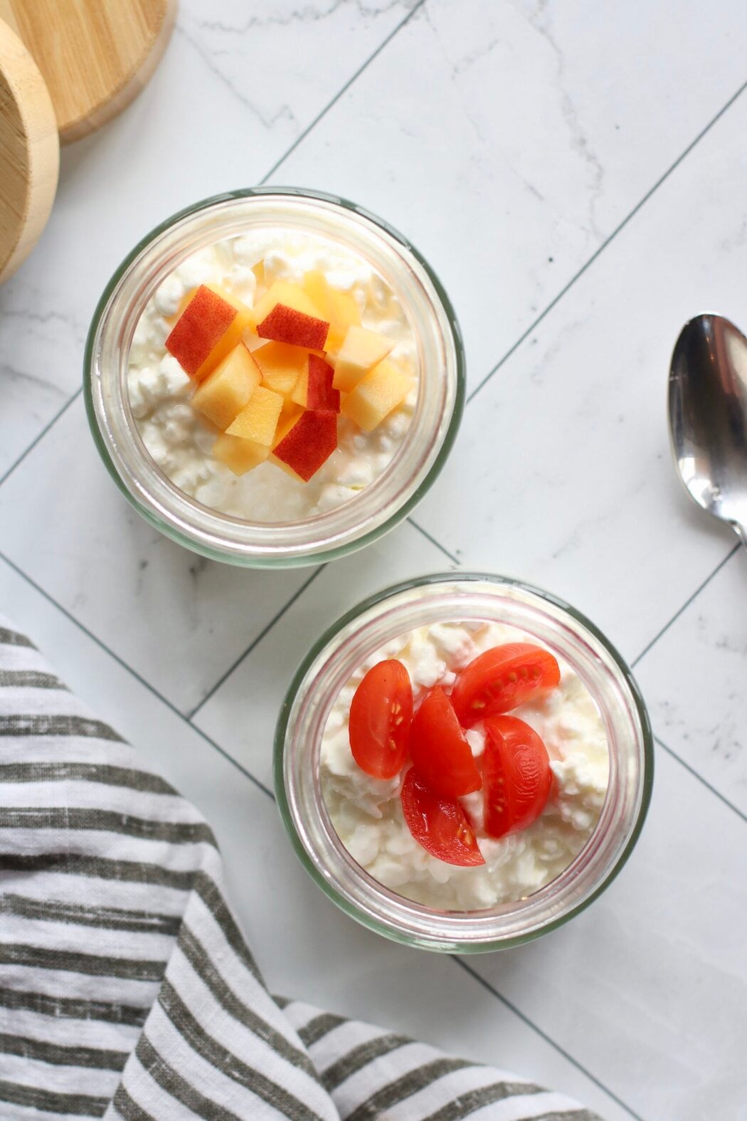 Overhead view of two jars of cottage cheese topped with sliced peaches and sliced cherry tomatoes. 
