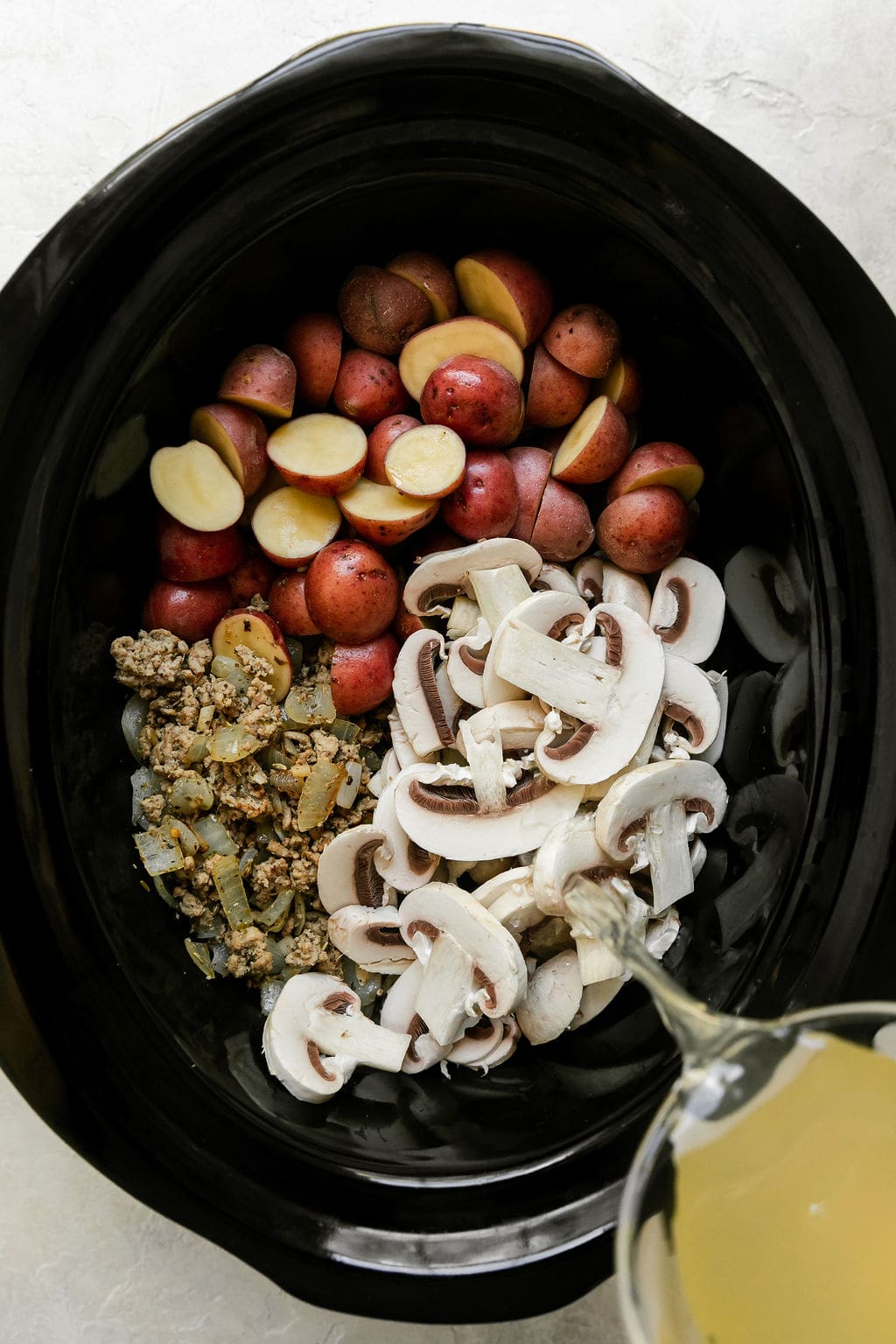 Broth being poured into black crockpot filled with all ingredients for zuppa toscana soup.