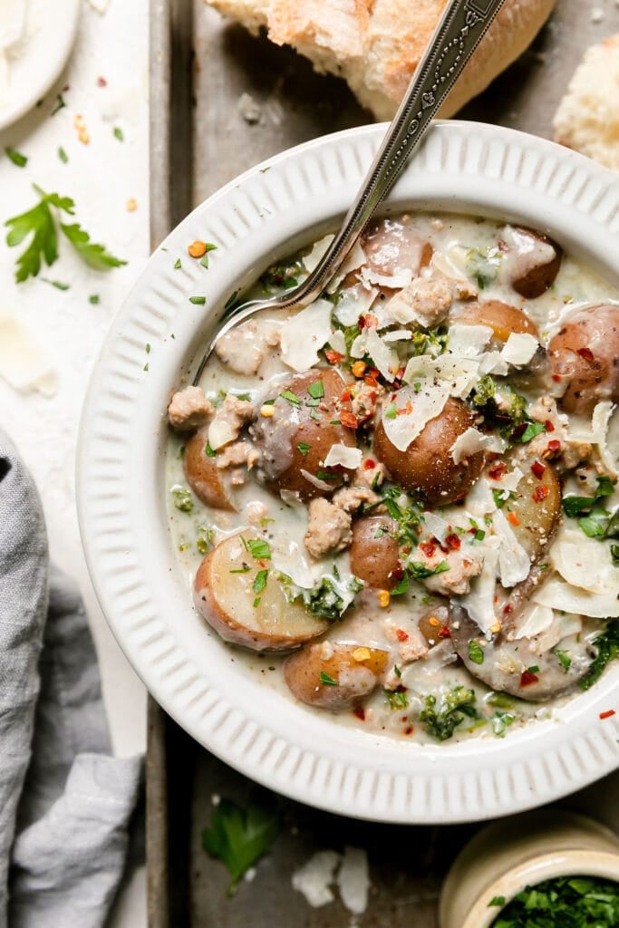 Overhead view white bowl filled with crockpot zuppa toscana topped with shaved Parmesan cheese, placed on silver baking sheet with crusty bread in background.