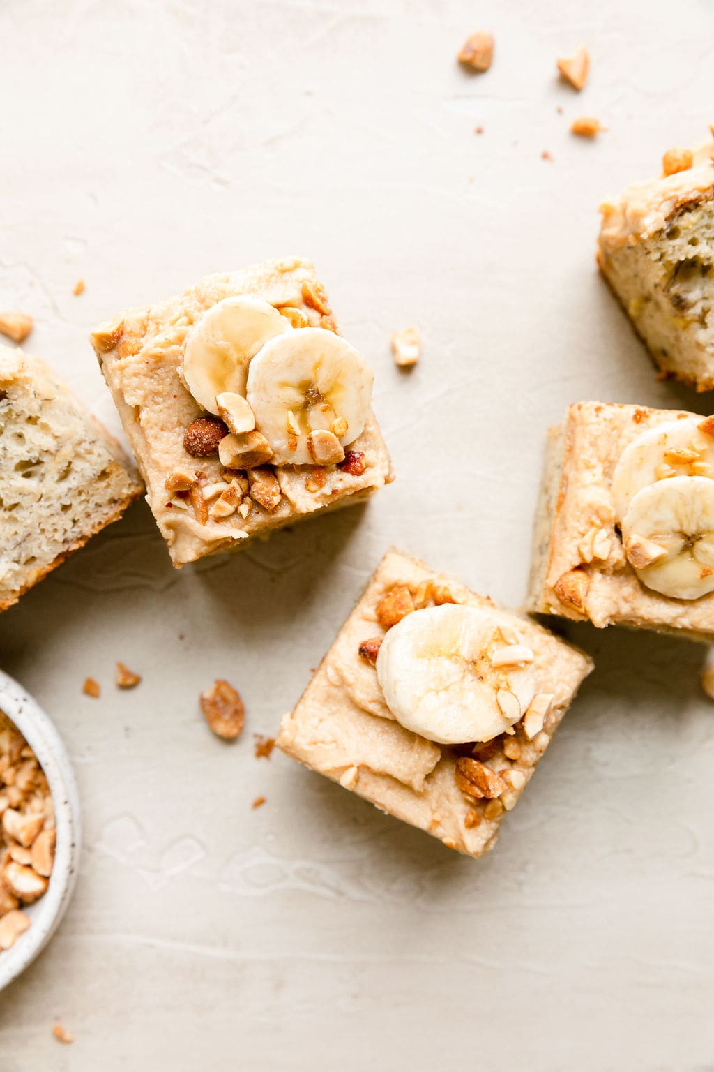Overhead view several pieces banana cake topped with frosting and banana slices arranged on white countertop.