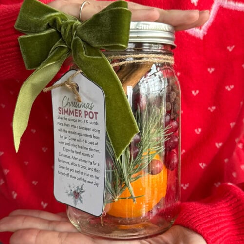 A woman in a red holiday sweater holding a glass mason jar filled with cranberries, orange, and cinnamon sticks. The jar has a pretty gift tag attached to it.