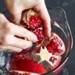 Pomegranate sections with arils in a bowl of water, two hands working to remove pomegranate arils from membranes of pomegranate.
