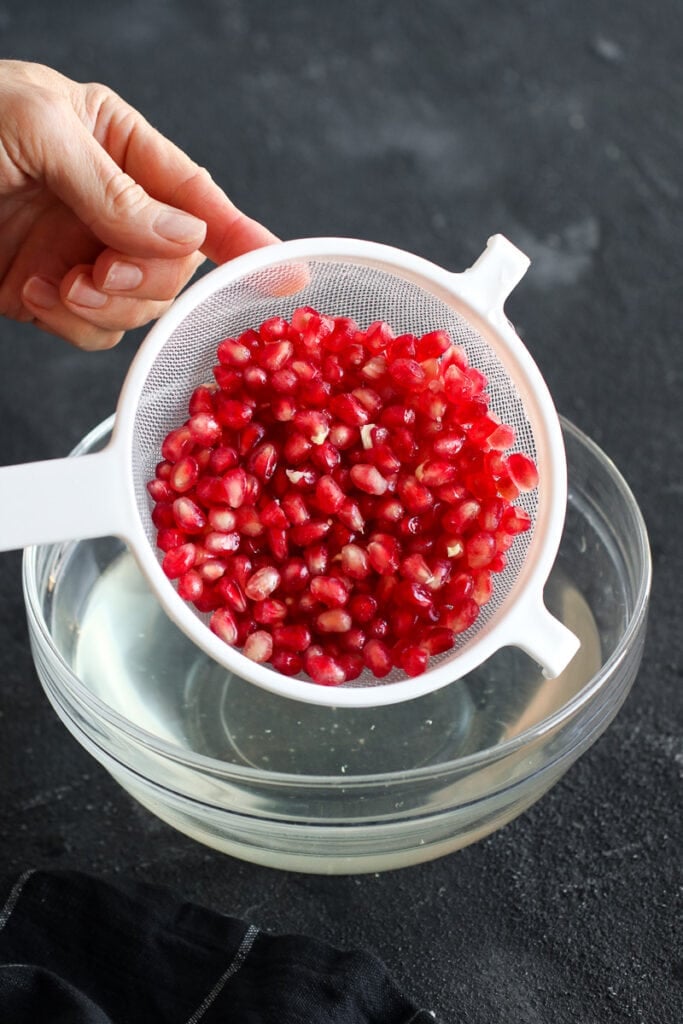 Pomegranate arils in a white colander over a bowl of water from being strained.