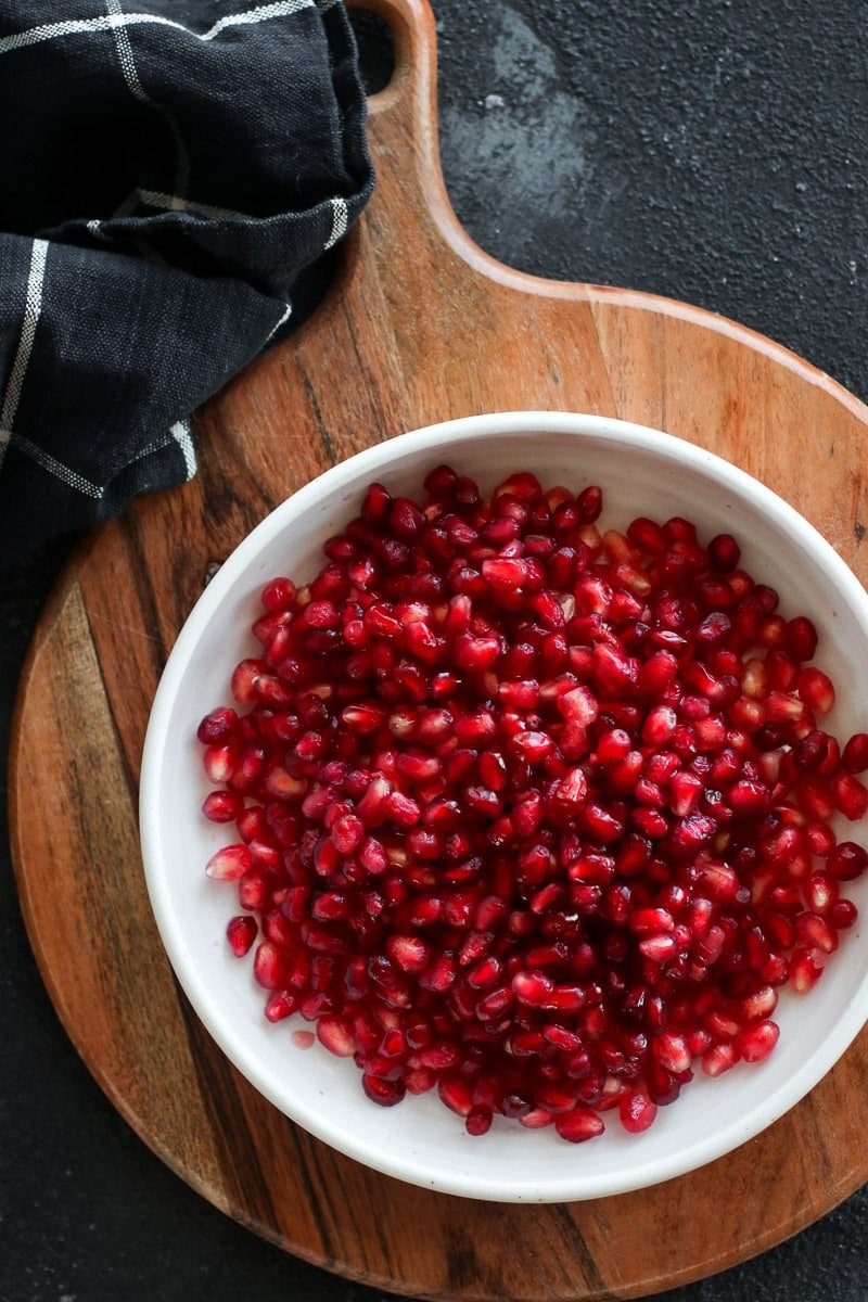 A white bowl filled with pomegranate arils on a round wooden cutting board.