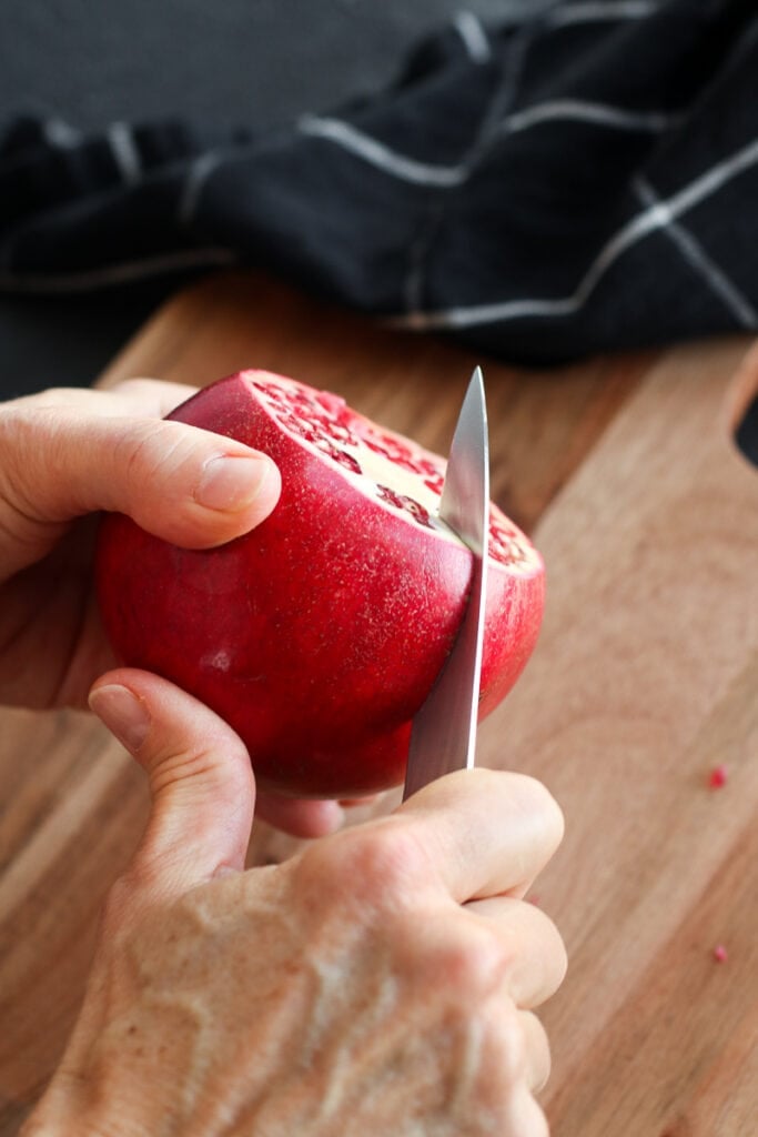 A pomegranate being held in one hand, other hand using pairing knife to cut down sides. 