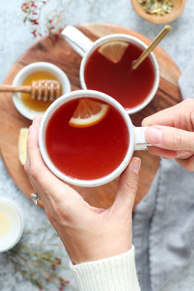 Two hands holding a mug of Medicine Ball Tea over a wooden tray holding another mug of tea.