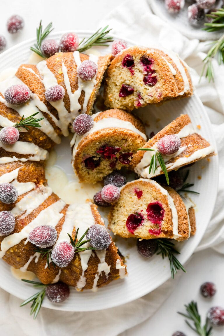 Overhead view of a sliced bundt cranberry orange olive oil cake topped with sugared cranberries and fresh rosemary sprigs.