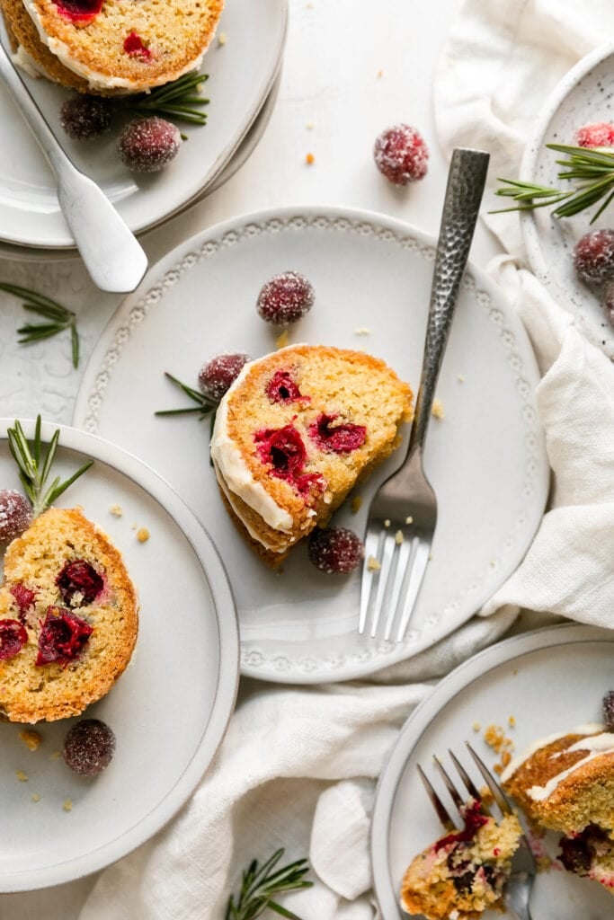 Slices of cranberry orange olive oil bundt cake on white plate topped with white icing, sugared cranberries and rosemary sprigs.