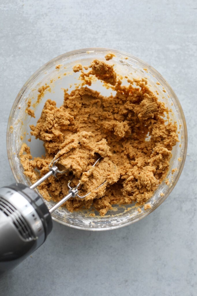 Peanut butter blossoms cookie dough in clear glass mixing bowl with hand mixer beaters in dough.