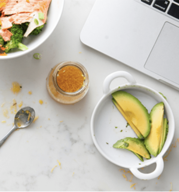 Corner of silver laptop on counter with plate with avocado slices and small jar of salad dressing.