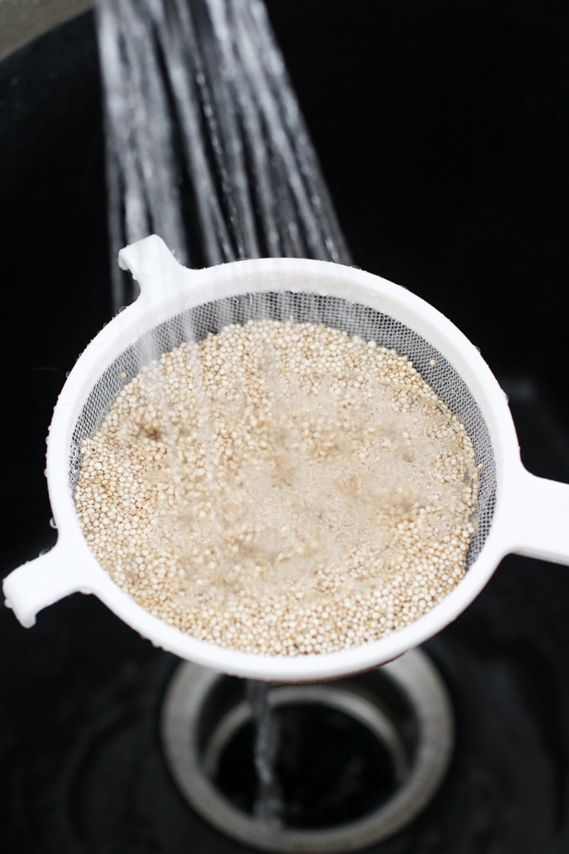 A small mesh strainer filled with uncooked quinoa, water spraying into strainer to rinse quinoa
