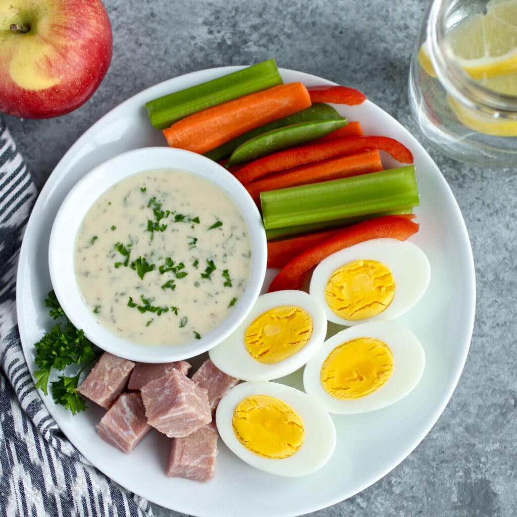 Overhead view of a plate of vegetable sticks, hard boiled eggs, diced ham and a ranch dip. 