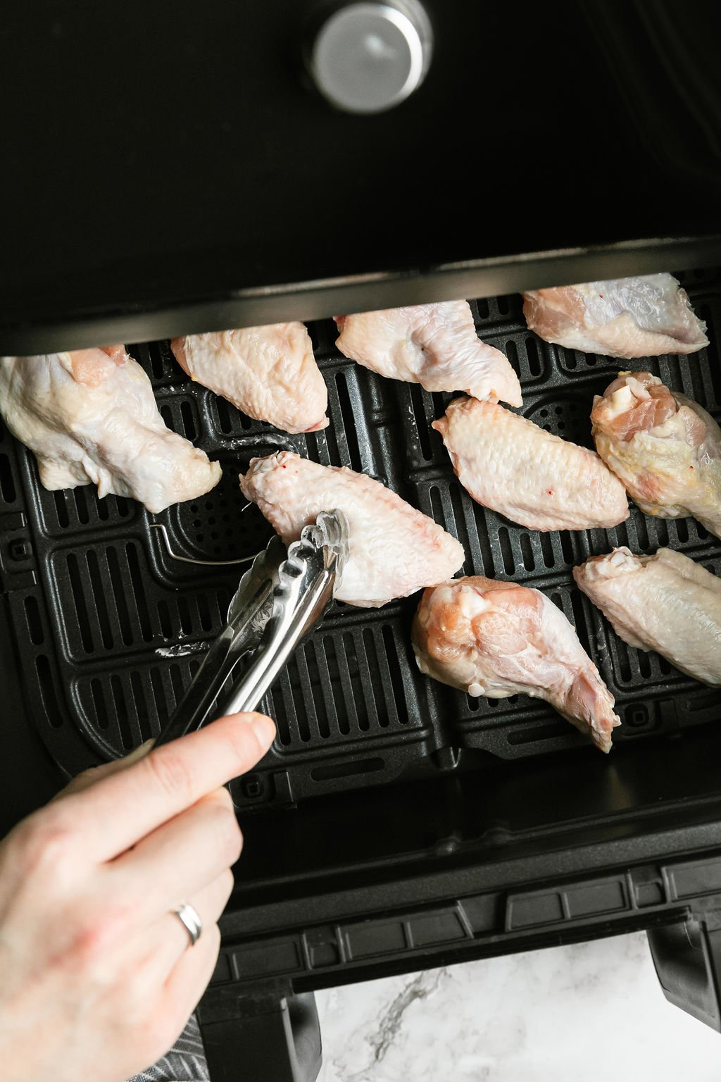Raw chicken wings being placed in air fryer basket with tongs.