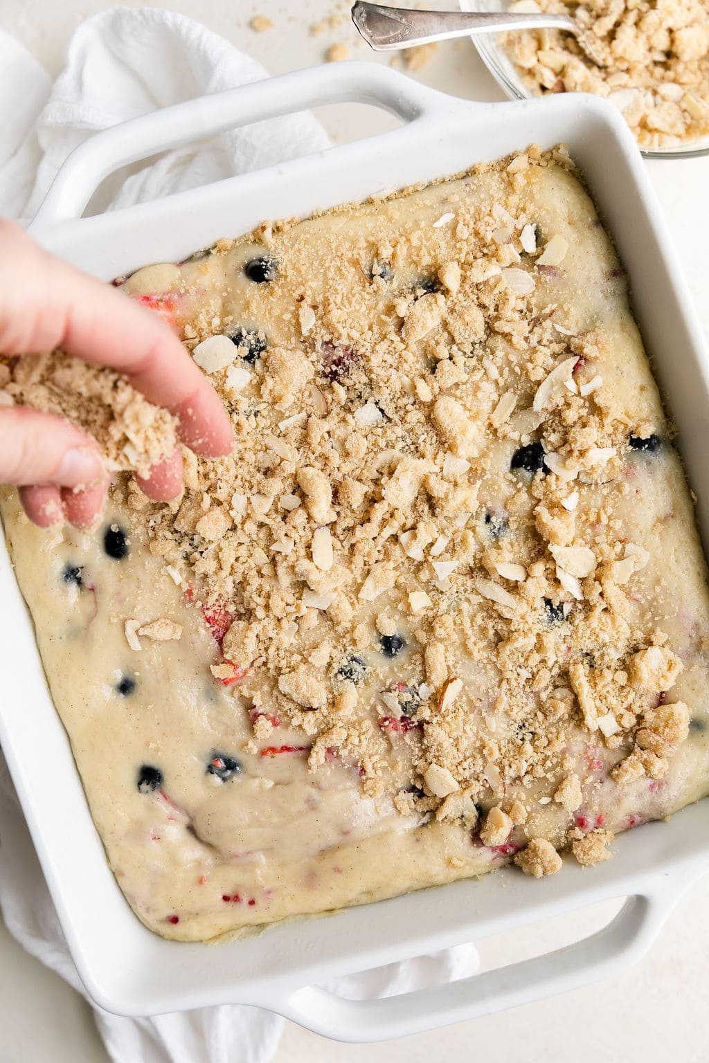 Overhead view white baking dish with mixed berry breakfast cake batter, crumb topping being sprinkled on top.