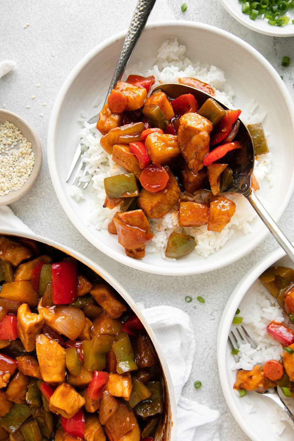 Spoonful of sweet and sour chicken being placed over bowl of rice, skillet of sweet and sour chicken in bottom left.