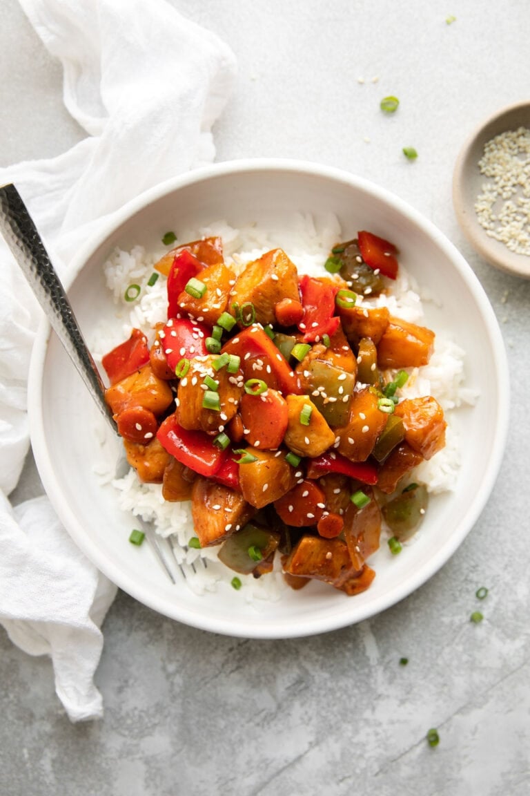 Overhead view of a plate of white rice topped with sweet and sour chicken and sprinkled with sesame seeds and green onions. 