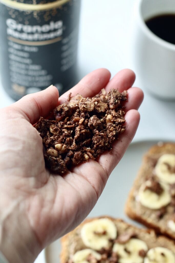 A hand holding serving of Struesli grain-free granola over peanut butter banana toast.