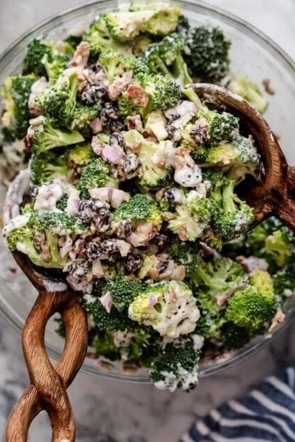 Overhead view of a full recipe of broccoli salad in a clear serving bowl with a wooden serving spoon in the bowl. 
