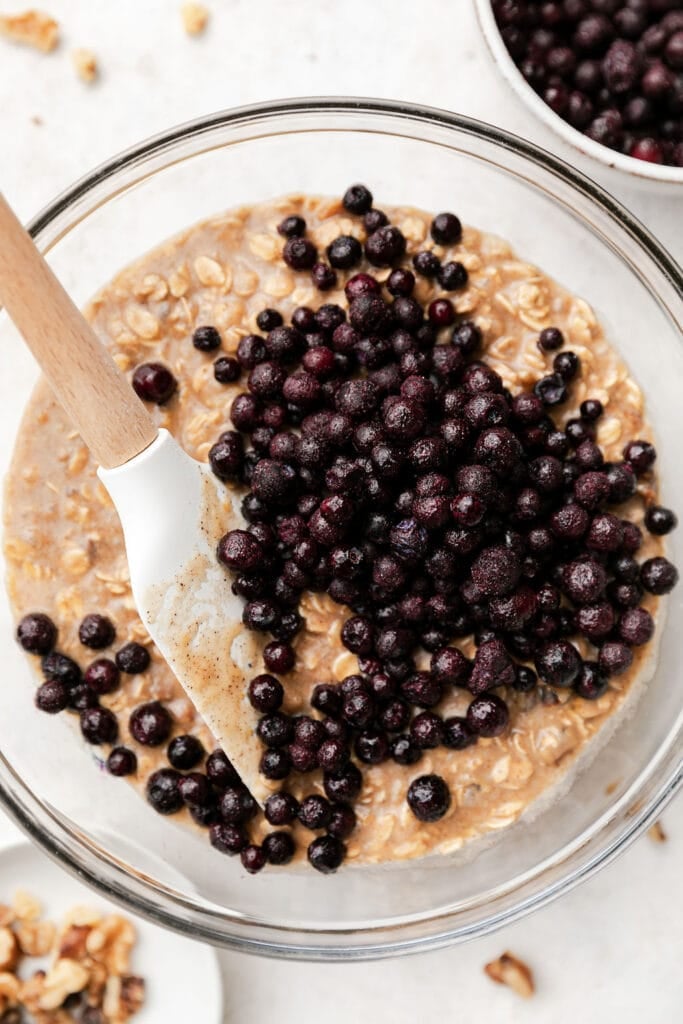 Frozen wild blueberries being folded into oatmeal breakfast bar batter in clear glass mixing bowl