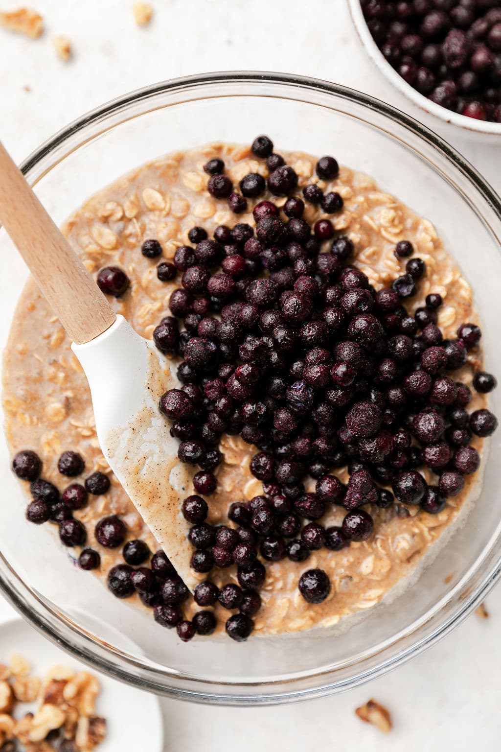 Frozen wild blueberries being folded into oatmeal breakfast bar batter in clear glass mixing bowl.
