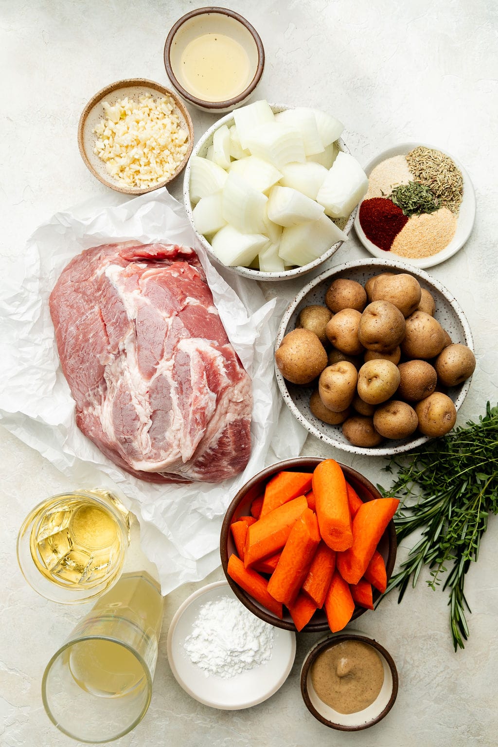The ingredients that will be used to make crock pot pork roast, including pork shoulder, potatoes, carrots, spices, onions, minced garlic, Dijon mustard, broth, and white wine. 