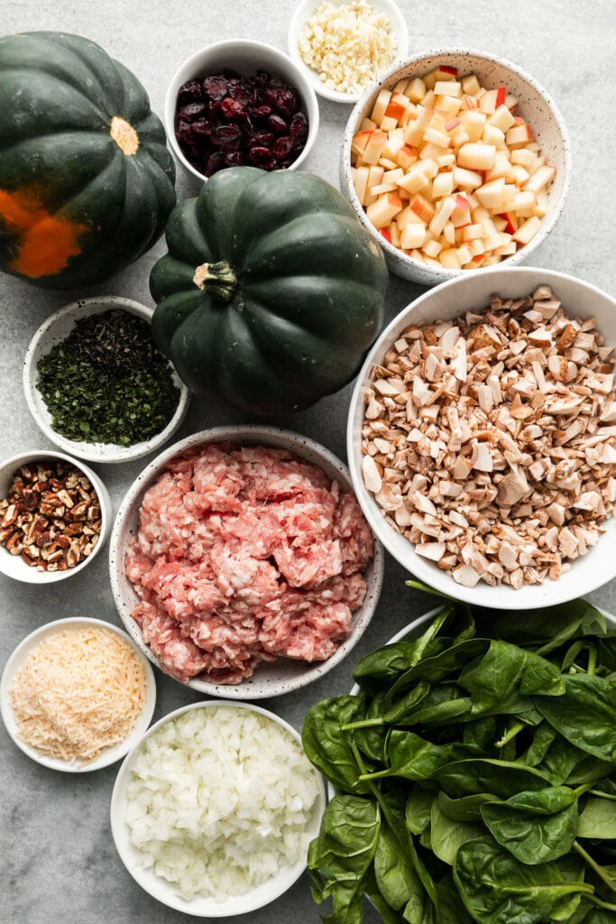 All ingredients for sausage stuffed acorn squash arranged together in small bowls with two acorn squash in the middle of them