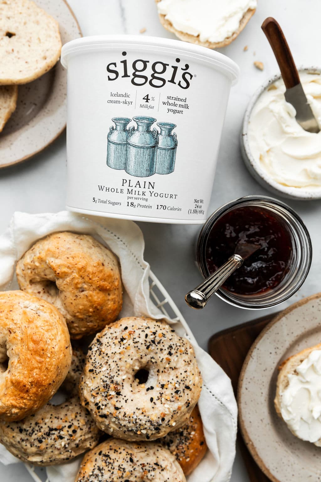 An overhead view of a counter with Greek yogurt bagels, a container of Siggi's plain 4% milkfat yogurt,  jam and cream cheese both with knives in the containers. 